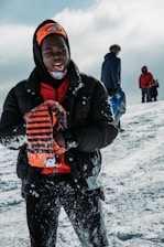 A person dressed in winter clothing, standing on a snowy surface and holding an orange and black striped object. They are wearing an orange hat with a logo on it. In the background, two other individuals are also dressed in winter clothing, one carrying a snow sled.