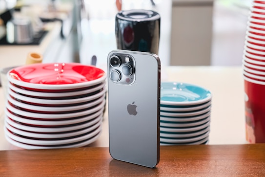 A sleek, modern smartphone is propped upright on a wooden surface, surrounded by neatly stacked colorful ceramic plates. The phone's metallic finish and prominent camera lenses are noticeable. The plates include stacks of red and teal against a softly blurred background, suggesting a casual and cozy setting.