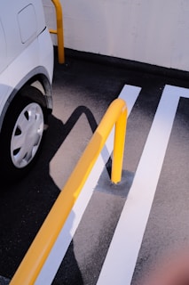 A section of a parking lot with a white vehicle parked next to yellow barrier poles. The surface is asphalt with white lines marking the parking area. The sunlight casts shadows of the poles and vehicle on the ground.