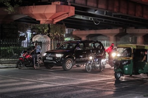 A nighttime urban street scene featuring a black SUV, several motorcycles, and a green auto-rickshaw. The vehicles are stopped near an intersection under a bridge, with illuminated headlights creating reflections on the wet road surface. A few people can be seen around the vehicles, including one wearing a reflective vest.