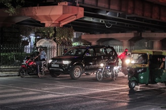A nighttime urban street scene featuring a black SUV, several motorcycles, and a green auto-rickshaw. The vehicles are stopped near an intersection under a bridge, with illuminated headlights creating reflections on the wet road surface. A few people can be seen around the vehicles, including one wearing a reflective vest.