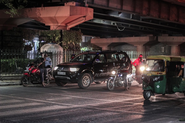 A nighttime urban street scene featuring a black SUV, several motorcycles, and a green auto-rickshaw. The vehicles are stopped near an intersection under a bridge, with illuminated headlights creating reflections on the wet road surface. A few people can be seen around the vehicles, including one wearing a reflective vest.