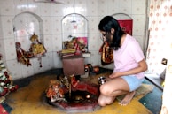 A person is kneeling in a small shrine or prayer room, holding a metal container. The walls are tiled and contain recessed shelves holding various religious idols and decorations. A small flame or diya is visible in front of the person.