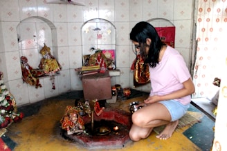 A person is kneeling in a small shrine or prayer room, holding a metal container. The walls are tiled and contain recessed shelves holding various religious idols and decorations. A small flame or diya is visible in front of the person.