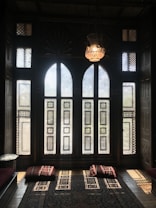 An intricately decorated room featuring large, arched windows with patterned screens that allow sunlight to filter through, creating geometric shadows on the floor. A hanging ornate lamp complements the traditional, rustic ambiance. Cushions with striped patterns are placed on a richly designed carpet.