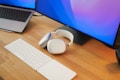 A modern workspace setup featuring a laptop partially visible with a wooden table surface. Nearby, a pair of white over-ear headphones rests next to a wireless keyboard and trackpad. A large monitor displays a pastel gradient, adding a touch of color to the scene.