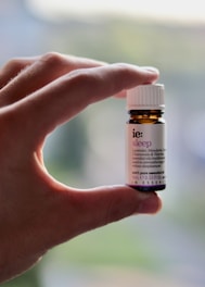 A hand is holding a small amber-colored bottle with a white label and cap. The label has text on it, indicating it is an essential oil named 'sleep' with ingredients like lavender, mandarin, chamomile, and valerian.