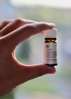 A hand is holding a small amber-colored bottle with a white label and cap. The label has text on it, indicating it is an essential oil named 'sleep' with ingredients like lavender, mandarin, chamomile, and valerian.