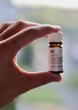 A hand is holding a small amber-colored bottle with a white label and cap. The label has text on it, indicating it is an essential oil named 'sleep' with ingredients like lavender, mandarin, chamomile, and valerian.