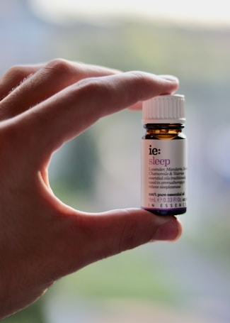 A hand is holding a small amber-colored bottle with a white label and cap. The label has text on it, indicating it is an essential oil named 'sleep' with ingredients like lavender, mandarin, chamomile, and valerian.