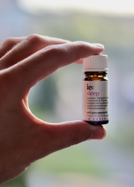 A hand is holding a small amber-colored bottle with a white label and cap. The label has text on it, indicating it is an essential oil named 'sleep' with ingredients like lavender, mandarin, chamomile, and valerian.