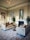 Luxurious living area featuring elegant velvet sofas with tufted designs and ornamental pillows. A round wooden table is positioned between the sofas. A grand, intricate chandelier hangs from a ceiling adorned with decorative molding. The room is decorated with framed artwork and a large mirror above the fireplace. Plants are placed on either side of the fireplace, enhancing the overall aesthetic.