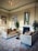 Luxurious living area featuring elegant velvet sofas with tufted designs and ornamental pillows. A round wooden table is positioned between the sofas. A grand, intricate chandelier hangs from a ceiling adorned with decorative molding. The room is decorated with framed artwork and a large mirror above the fireplace. Plants are placed on either side of the fireplace, enhancing the overall aesthetic.