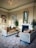 Luxurious living area featuring elegant velvet sofas with tufted designs and ornamental pillows. A round wooden table is positioned between the sofas. A grand, intricate chandelier hangs from a ceiling adorned with decorative molding. The room is decorated with framed artwork and a large mirror above the fireplace. Plants are placed on either side of the fireplace, enhancing the overall aesthetic.