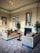 Luxurious living area featuring elegant velvet sofas with tufted designs and ornamental pillows. A round wooden table is positioned between the sofas. A grand, intricate chandelier hangs from a ceiling adorned with decorative molding. The room is decorated with framed artwork and a large mirror above the fireplace. Plants are placed on either side of the fireplace, enhancing the overall aesthetic.