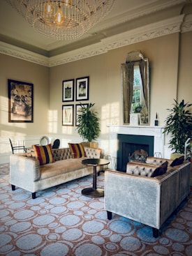 Luxurious living area featuring elegant velvet sofas with tufted designs and ornamental pillows. A round wooden table is positioned between the sofas. A grand, intricate chandelier hangs from a ceiling adorned with decorative molding. The room is decorated with framed artwork and a large mirror above the fireplace. Plants are placed on either side of the fireplace, enhancing the overall aesthetic.