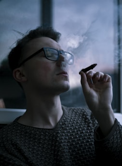 A person wearing glasses is indoors, holding a cigar or similar object near their mouth. Smoke gently rises from the cigar, partially obscuring the face. The background is dimly lit and features a window.