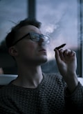 A person wearing glasses is indoors, holding a cigar or similar object near their mouth. Smoke gently rises from the cigar, partially obscuring the face. The background is dimly lit and features a window.