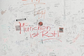 A wall filled with various handwritten notes, doodles, and stickers. The most prominent text is 'München ist Rot!' written in bold red letters. The wall also contains multiple scribbles, drawings, names, and dates in different handwriting styles and colors.