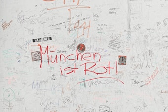 A wall filled with various handwritten notes, doodles, and stickers. The most prominent text is 'M&uuml;nchen ist Rot!' written in bold red letters. The wall also contains multiple scribbles, drawings, names, and dates in different handwriting styles and colors.