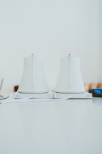 Two sleek, white electric kettles sit side by side on a light-colored countertop. They have a modern design with smooth surfaces and minimalistic features, and are positioned on their respective bases.