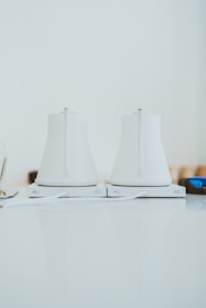 Two sleek, white electric kettles sit side by side on a light-colored countertop. They have a modern design with smooth surfaces and minimalistic features, and are positioned on their respective bases.