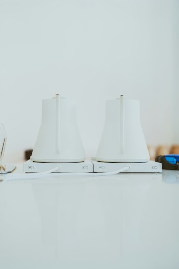 Two sleek, white electric kettles sit side by side on a light-colored countertop. They have a modern design with smooth surfaces and minimalistic features, and are positioned on their respective bases.