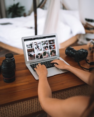 A person using a laptop on a wooden desk, surrounded by camera equipment, including lenses and a digital camera. The screen displays a gallery of photos in a grid layout. The blurred background reveals a cozy interior with a bed and plant.