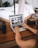 A person using a laptop on a wooden desk, surrounded by camera equipment, including lenses and a digital camera. The screen displays a gallery of photos in a grid layout. The blurred background reveals a cozy interior with a bed and plant.