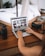 A person using a laptop on a wooden desk, surrounded by camera equipment, including lenses and a digital camera. The screen displays a gallery of photos in a grid layout. The blurred background reveals a cozy interior with a bed and plant.