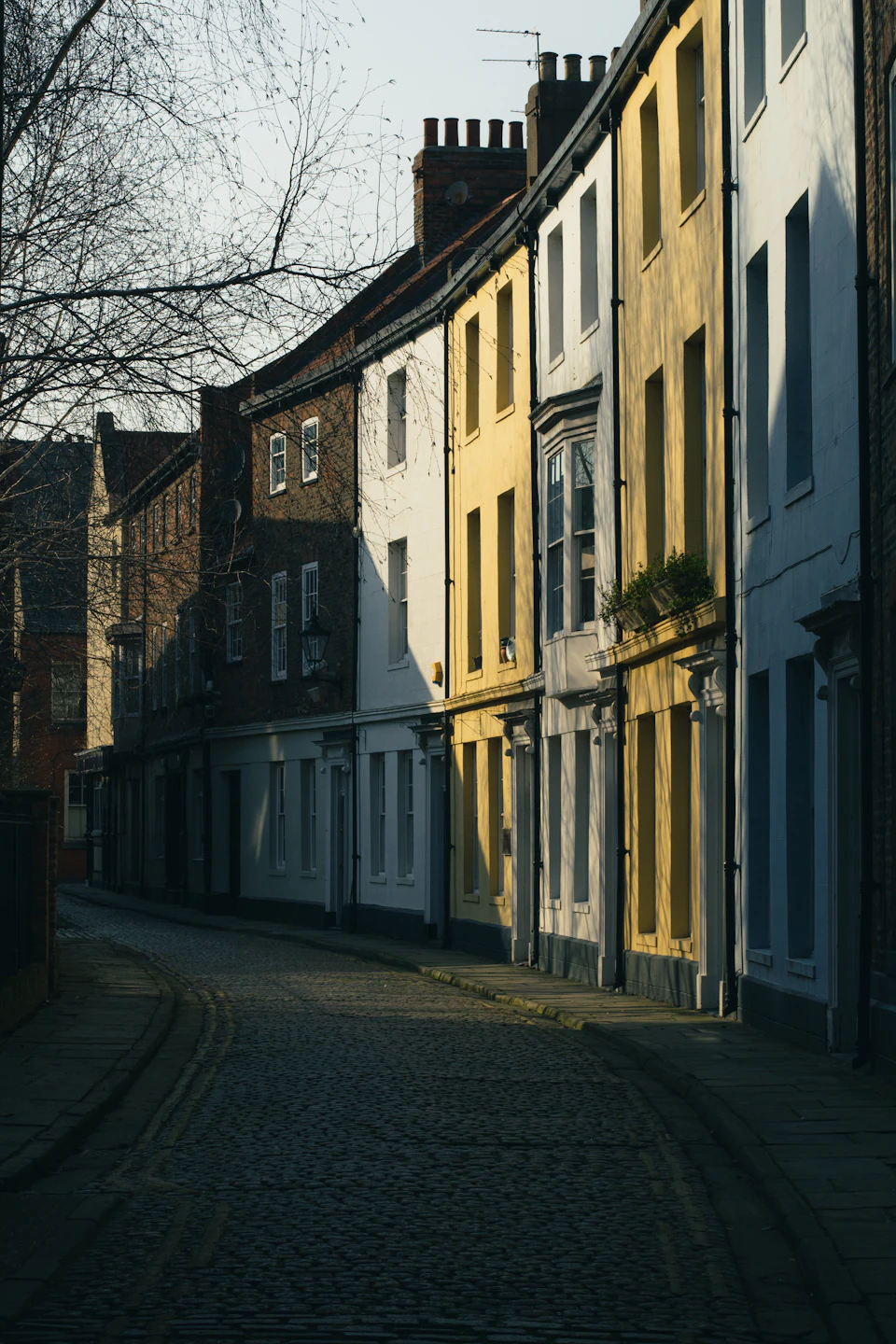 The image depicts a narrow cobblestone street lined with old, traditional brick buildings painted in shades of white and yellow. The buildings feature chimneys on their rooftops, adding to the rustic charm of the scene. A few trees can be seen along the sidewalk, providing some greenery amidst the stone structures. The perspective of the image is from ground level, looking down the street towards the horizon line, giving a sense of depth and distance.