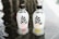 Two clear plastic bottles with black caps placed on a reflective surface. The bottles have a minimalist design featuring bold black text and checkmarks indicating '0 fat, 0 sugar, 0 calories'. Each bottle has a fruit image at the bottom representing different flavors.