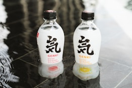 Two clear plastic bottles with black caps placed on a reflective surface. The bottles have a minimalist design featuring bold black text and checkmarks indicating '0 fat, 0 sugar, 0 calories'. Each bottle has a fruit image at the bottom representing different flavors.