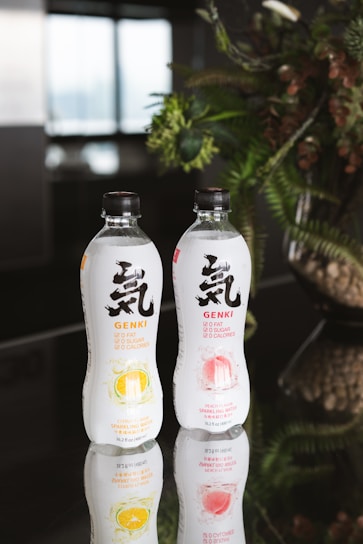 Two bottles of Genki sparkling water are placed on a reflective surface. The left bottle is citrus flavored, and the right one is peach flavored. Both bottles have a simple design with bold Japanese characters and are set against a softly blurred background of green leafy plants and a modern interior.