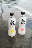 Two bottles of Genki sparkling water are placed on a gray stone surface. The bottle on the left has a citrus lemon flavor, depicted by an image of a lemon slice, while the bottle on the right features a peach flavor, illustrated by a peach image. The bottles are transparent with black caps and display bold black and red text.