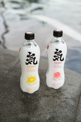 Two bottles of Genki sparkling water are placed on a gray stone surface. The bottle on the left has a citrus lemon flavor, depicted by an image of a lemon slice, while the bottle on the right features a peach flavor, illustrated by a peach image. The bottles are transparent with black caps and display bold black and red text.