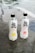 Two bottles of Genki sparkling water are placed on a gray stone surface. The bottle on the left has a citrus lemon flavor, depicted by an image of a lemon slice, while the bottle on the right features a peach flavor, illustrated by a peach image. The bottles are transparent with black caps and display bold black and red text.