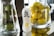 A bottle of Genki citrus sparkling water is placed on a reflective surface, flanked by two large glass containers. One container holds green avocados, and the other is filled with bright yellow lemons. The setting appears to be well-lit, with a focus on the freshness and vibrancy of the fruits.