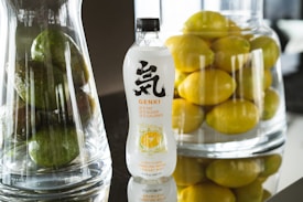 A bottle of Genki citrus sparkling water is placed on a reflective surface, flanked by two large glass containers. One container holds green avocados, and the other is filled with bright yellow lemons. The setting appears to be well-lit, with a focus on the freshness and vibrancy of the fruits.
