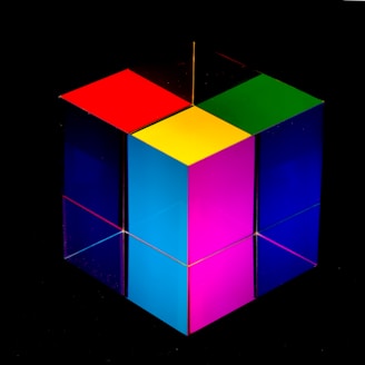 A colorful, transparent cube with each face displaying a different vibrant color, such as red, green, yellow, pink, and blue. The cube appears to be made of glass or a similar material, reflecting light and showcasing a glossy finish. It is set against a solid black background, which enhances the vividness of the colors.