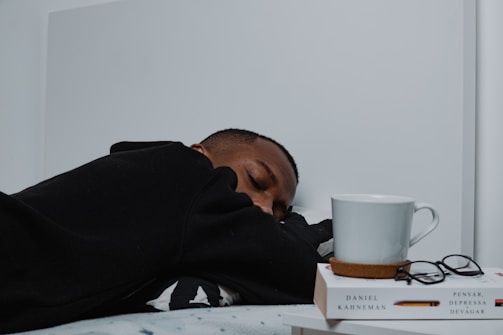 A person in a black hoodie is resting with their head on a surface. Next to them is a white mug placed on two stacked books, alongside a pair of eyeglasses. The books, one of which is authored by Daniel Kahneman, are positioned neatly and complement the minimalist setting.