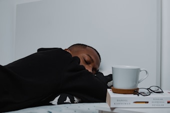 A person in a black hoodie is resting with their head on a surface. Next to them is a white mug placed on two stacked books, alongside a pair of eyeglasses. The books, one of which is authored by Daniel Kahneman, are positioned neatly and complement the minimalist setting.
