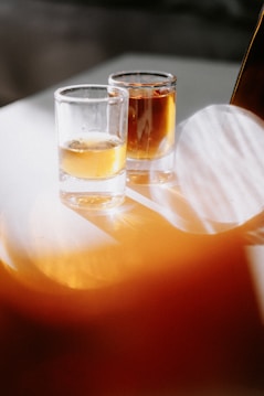 Two shot glasses filled with an amber-colored liquid are placed on a surface. The lighting casts soft shadows and reflections, creating a warm and inviting atmosphere.
