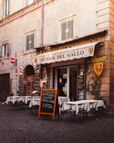 A quaint Italian restaurant with an outdoor seating area displaying white tablecloths covering round tables. The building has a rustic appearance with weathered stone walls and shuttered windows. A chalkboard menu is placed outside, advertising various pasta dishes and pizza. Ivy climbs part of the wall beside a yellow and red emblem of a rooster.