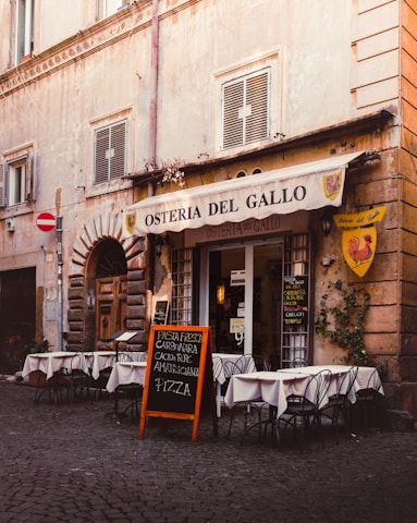 A quaint Italian restaurant with an outdoor seating area displaying white tablecloths covering round tables. The building has a rustic appearance with weathered stone walls and shuttered windows. A chalkboard menu is placed outside, advertising various pasta dishes and pizza. Ivy climbs part of the wall beside a yellow and red emblem of a rooster.