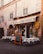 A quaint Italian restaurant with an outdoor seating area displaying white tablecloths covering round tables. The building has a rustic appearance with weathered stone walls and shuttered windows. A chalkboard menu is placed outside, advertising various pasta dishes and pizza. Ivy climbs part of the wall beside a yellow and red emblem of a rooster.