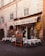 A quaint Italian restaurant with an outdoor seating area displaying white tablecloths covering round tables. The building has a rustic appearance with weathered stone walls and shuttered windows. A chalkboard menu is placed outside, advertising various pasta dishes and pizza. Ivy climbs part of the wall beside a yellow and red emblem of a rooster.