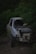 A damaged car with its front partially smashed and windows broken is parked amidst dense green foliage. The car's engine and other internal components are exposed, indicating significant wreckage. The vegetation surrounding the vehicle gives a wilderness-like feel.