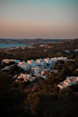 A hillside covered with white, modern-style buildings is surrounded by lush greenery. The buildings are bathed in warm, golden light, suggesting either dawn or dusk. The calm sea extends into the background while hills covered in trees fill the foreground and distance.