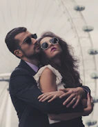a man in a suit and sunglasses kissing a woman in front of a ferris wheel